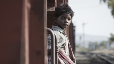 Saroo (Sunny Pawar) står på trappsteget till ett tåg och ser rakt in i kameran.