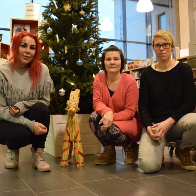 Tre kvinnor sitter på huk framför en pyntad julgran i Kimitos bibliotek.