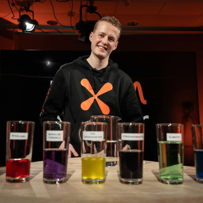 Redaktör Niklas Grönholm står i studion framför en kanna vatten och åtta olika glas.