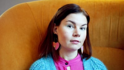 Emilia Kujala istuu oranssinkeltaisella nojatuolilla, pitää käsiä jalkojensa päällä ja katsoo suoraan kameraan.