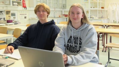 En ung man och en ung kvinna sitter i ett i övrigt tomt klassrum. De har en öppen dator framför sig.