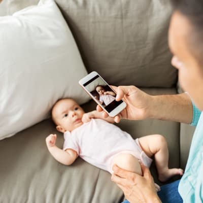 En man tar en bild med sin telefon på ett spädbarn som ligger i soffan.