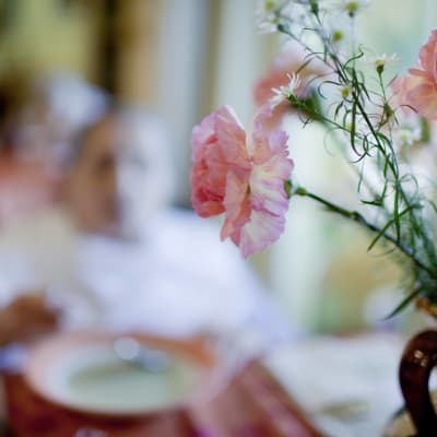 I förgrunden syns en bukett med blommor på ett bord, i bakgrunden syns en äldre kvinna på ett äldre boende som äter soppa.