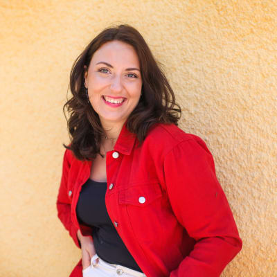 Sv Yle Barns reporter Charlotte Lindberg står med en röd jeansjacka mot en gul vägg och ler. 