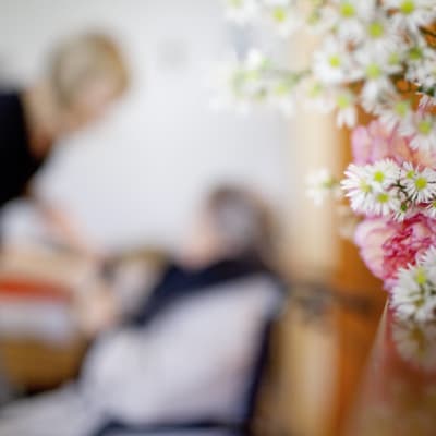 I förgrunden syns en bukett med blommor som står på ett bord. I bakgrunden syns en åldring som får hjälp av en yngre person, anställd, på ett äldreboende.