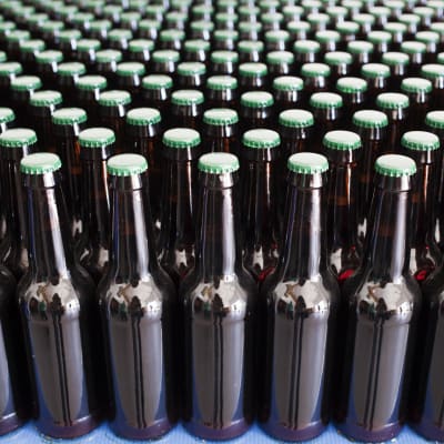 Närbild på en mängd mörkbruna ölflaskor med grön kapsyl som står på ett blått underlag.