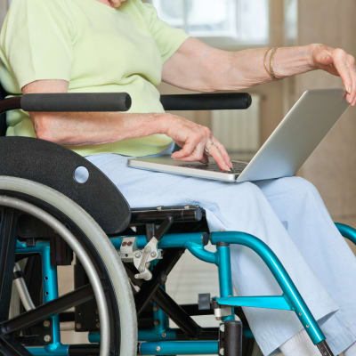 En äldre person sitter med bärbar dator i famnen i en rullstol.
