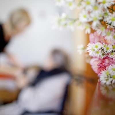 I förgrunden syns en bukett med blommor som står på ett bord. I bakgrunden syns en åldring som får hjälp av en yngre person, anställd, på ett äldreboende.