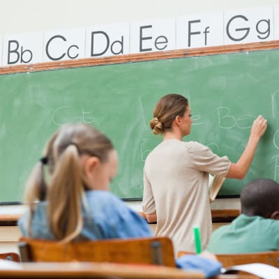 I förgrunden elever som sitter vid pulpeter, i bakgrunden en lärare som skriver på en tavla.