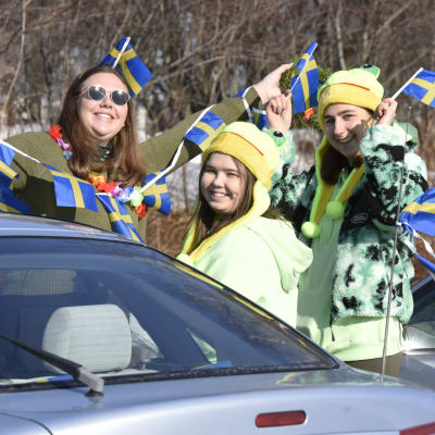 Tre kvinnor står intill en bil. De håller upp en rad med svenska flaggor.