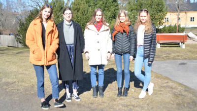 Fem unga kvinnor står framför kameran. Klädda i vinterrrockar på en skolgård. Ekenäs gymnasium. 