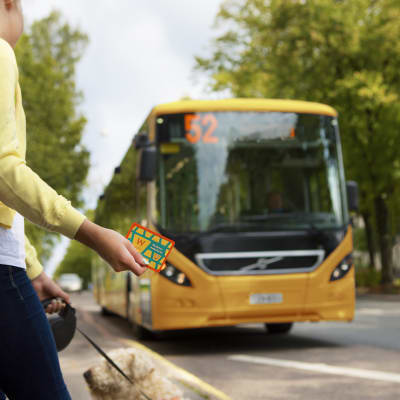 En flicka har ett busskort i handen och väntar på en gul buss som har nummer 52.