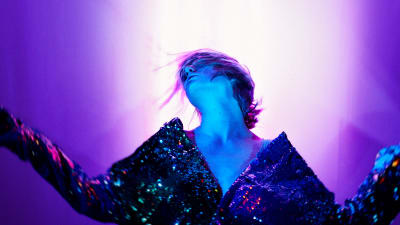 Skådespelaren Sue Lemström poserar med armarna utsträckta och huvudet bakåtlutat mot en neonviolett bakgrund.