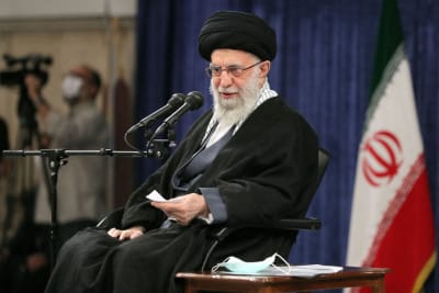 Irans högsta ledare Ali Khamenei ser välvillig ut på en officiellt sanktionerad bild. Han sitter med några mikrofoner framför sig och i bakgrunden finns Irans flagga.