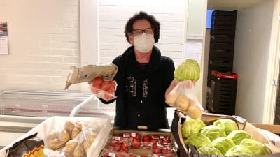 En kvinna med munskydd står bakom ett bord med matvaror på Hon håller tomater, potatis, kål och bröd i händerna.