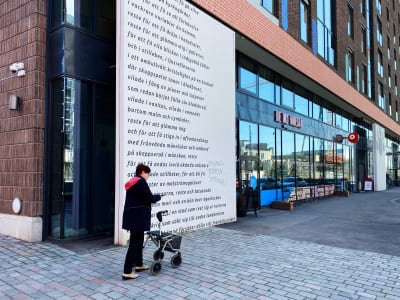 En kvinna med rollator går framför ett bostadshus och en vägg fylld med text på svenska.