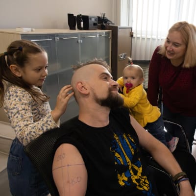 Miehen tyttäret koskettavat isän päätä, isä hymyilee. Äiti katsoo lapsia ja hymyilee.
