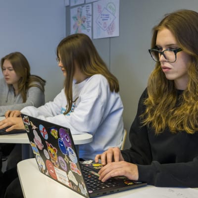 Tre elever sitter vid sina pulpeter i ett klassrum och arbetar på bärbara datorer. 