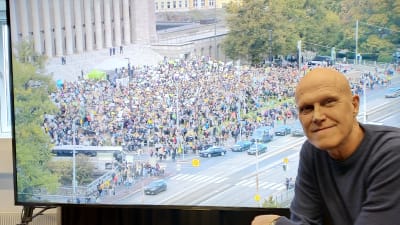 Thomas Wallgren i Svenska Yles lokaler framför en tv-skärm. På tv-bilden syns klimatdemonstranter utanför riksdagen den 27 september.
