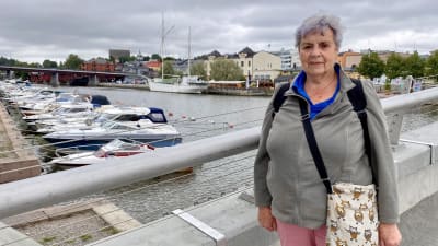 En äldre dam står på gångbron i Borgå och tittar in i kameran. Bakom henne syns Borgå å.