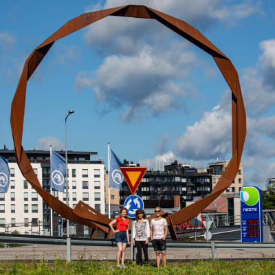 Tre personer poserar framför en cirkelformad skulptur gjord av rostigt stål.