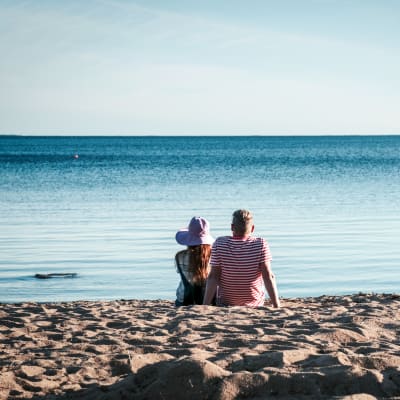 Hattupäinen nainen ja raitapaitainen mies istuvat hiekkarannalla katsomassa merta.