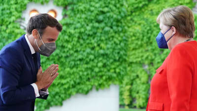 Emmanuel Macron och Angela Merkel står mittemot varandra. Macron gör en hälsning genom att buga och lägga ihop händerna med handflatorna mot varandra i brösthöjd.
