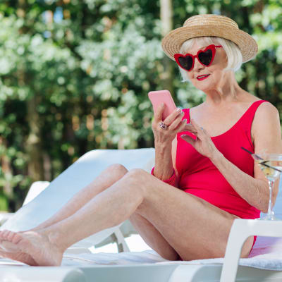 Äldre dam i baddräkt och solglasögon sitter på solstol och tittar leende på telefon 