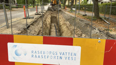 En gul grävmaskin vid en delvis uppgrävd gata. I förgrunden står det "Raseborgs vatten Raaseporin vesi" på en skylt. Hus och träd syns också.