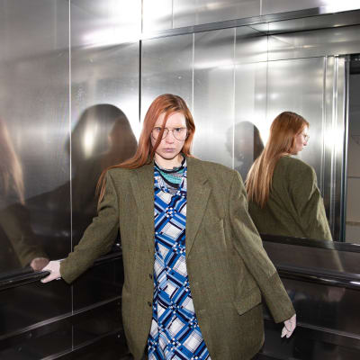 En ung kvinna står i en hiss och ser rakt in i kameran samtidigt som hon håller i hissräcket. 