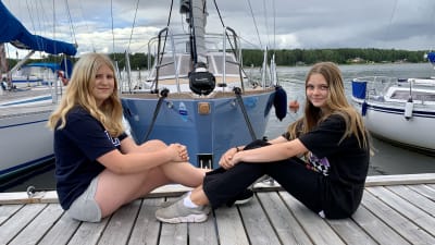Två unga flickor sitter mot varandra på en brygga framför en stor segelbåt i en hamn. 
