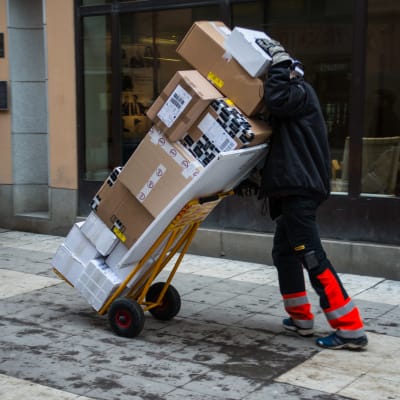 En person skuffar en bagagekärra med ett tiotal större paket på en snöig trottoar.