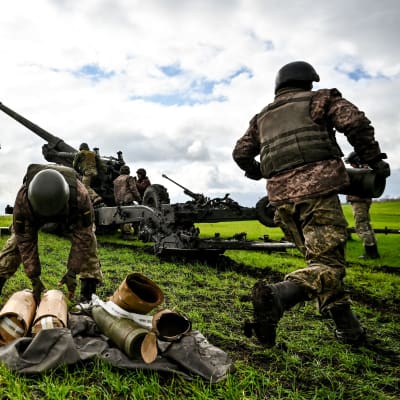 Ukrainalaissotilaat lataavat tykkiä, etualalla ammuksia, vihreä pelto. 