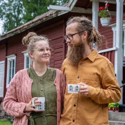 Ruut Luukkonen och Jakob Tjäder står vid ett gammalt rött trähus och dricker kaffe. 