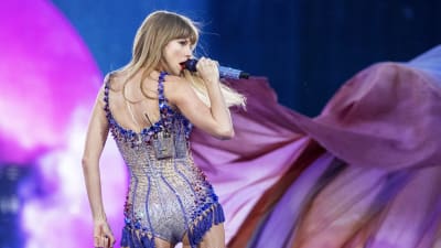 Taylor Swift står med ryggen mot kameran och sjunger in i en mikrofon på scenen. Hon har huvudet mot sidan så att hon tittar åt höger. Hon har på sig en glittrig dräkt.