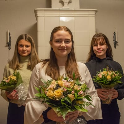 Tre kvinnor står i ett vardagsrum med blombuketter i händerna och tittar in i kameran.