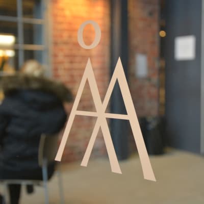 Åbo Akademis textlogo, ett Å och ett A klistrat på en glasdörr, insidan av huset med röd tegelvägg i bakgrunden.