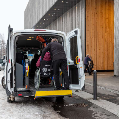 Taksinkuljettaja auttaa pyörätuolilla kulkevaa asiakasta taksin kyytiin Sairaala Novan pihassa.