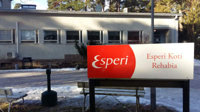 En rödvit skylt med texten Esperi Koti Rehabia framför en rappad beige byggnad i en våning.