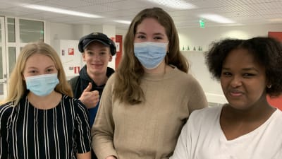 Fyra unga personer står intill varandra i skolmiljö, tittar in i kameran och ler. Två av dem bär munskydd.