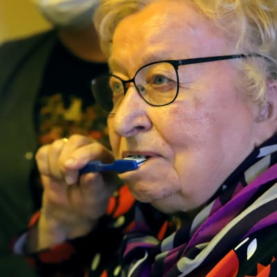 90-vuotias Eila Summanen pesee hampaitaan, sairaanhoitaja seuraa vieressä.