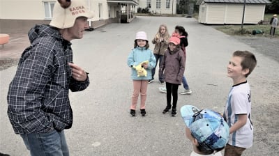 En man med bastuhatt står och talar med några barn på en skolgård. 