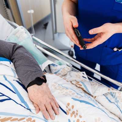 Närbild av en äldre persons hand och arm som ligger i en sjukhussäng. Hen har ett slags mätningsarmband på armen. En sjukskötare står bredvid sängen.