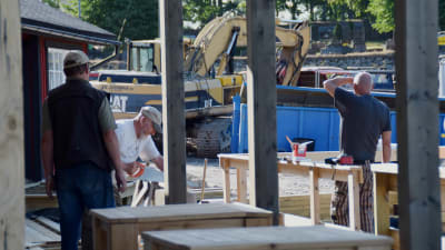 Byggarbetare jobbar vid ett bygge. En man sågar. I bakgrunden syns en grävmaskin.