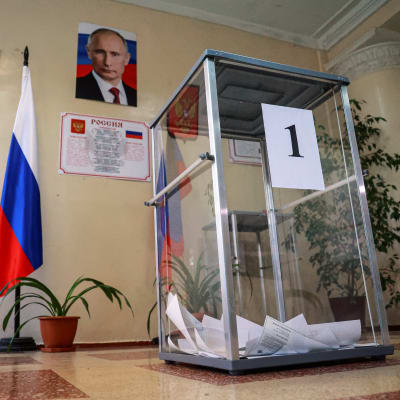 Lasiseinäinen vaaliuurna, jonka pohjalla on äänestyslipukkeita, seisoo lattialla äänestyspaikalla. Seinällä sen yläpuolella on presidentti Vladimir Putinin kuva. Putinin kuvan vieressä seisoo Venäjän lippu.