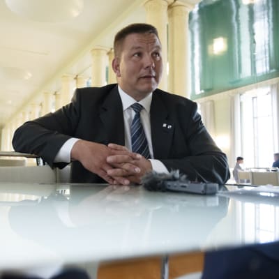 Riksdagsledamoten Juha Mäenpää (Sannf) intervjuas i riksdagen. Han har händerna i kors och han sitter vid ett bord och har en mikrofon framför sig.