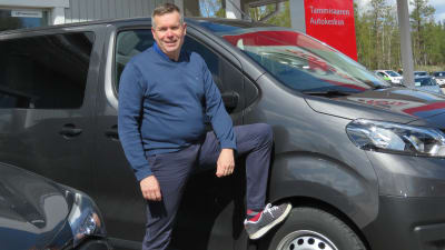 En man med kortklippt hår och blå tröja står utanför en bilaffär. Han står vid en bil med ena benet lyft och foten lutar på bilens framdäck.