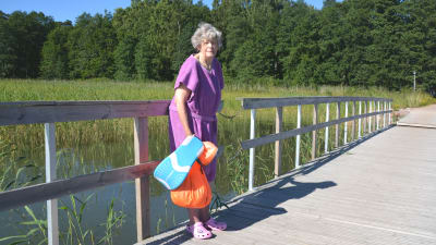 Äldre dam i badrock med simgrejer i handen står på bryggan vid Plagen