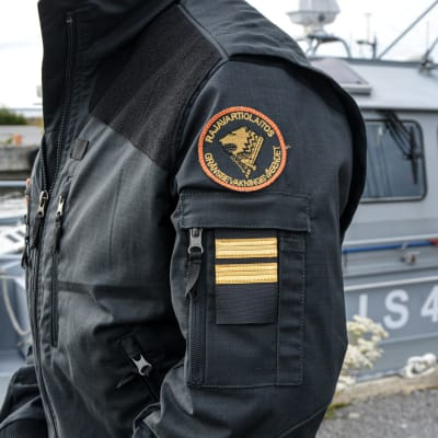 Anonym person i svart jacka. Gränsbevakningens emblem syns på axeln. En patrullbåt finns i bakgrunden.