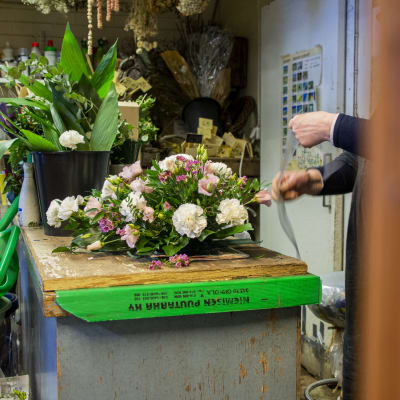 En blomsteruppsättning för en begravning ligger på ett bord i en blomsterhandel. Till höger syns en i personalen som håller på och gör en rosett.